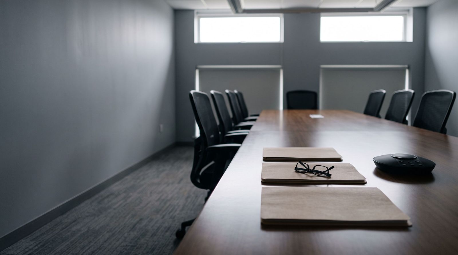 An empty boardroom table with three closed folders, a pair of reading glasses and a conference phone at one end.