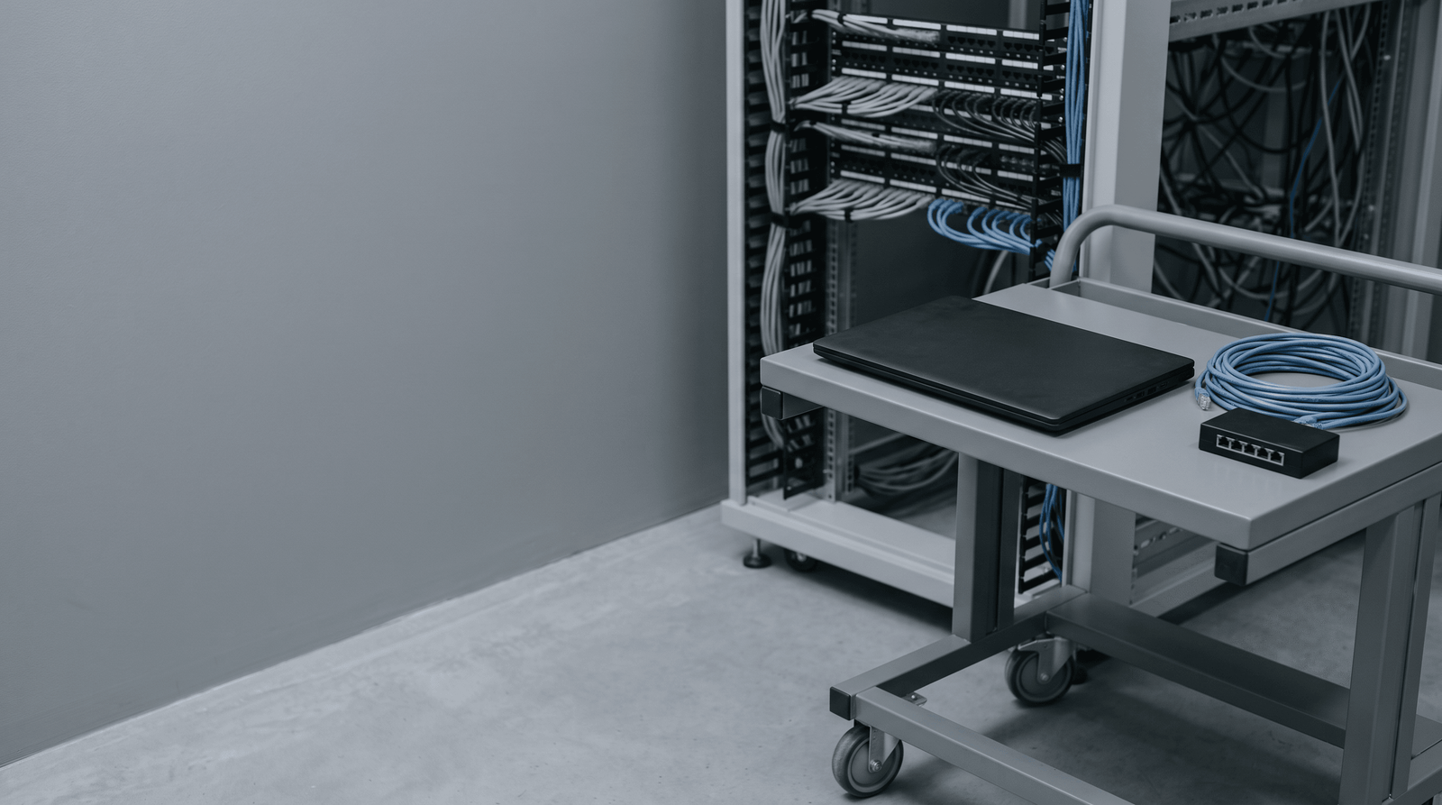 A closed laptop, a coiled blue ethernet cable and a small network tap on a technician's cart in front of an out-of-focus patch panel.