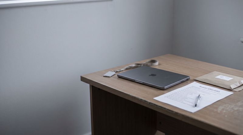 A returned company laptop closed on an office desk beside a staff lanyard and building fob, a ticked leaver checklist and a labelled envelope of keys.