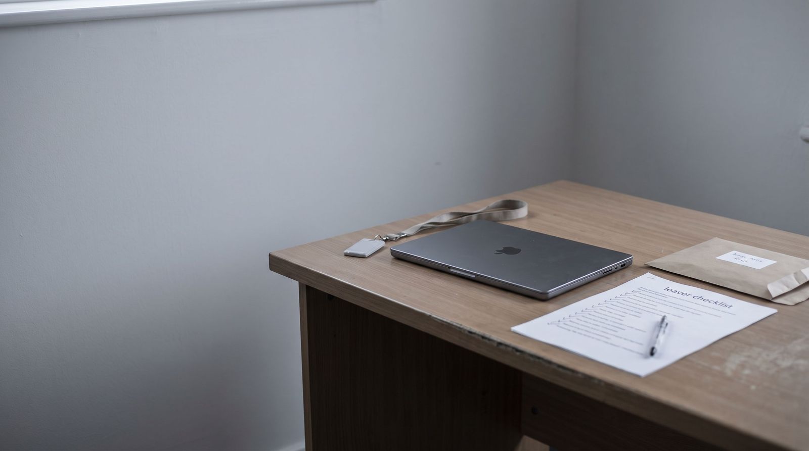 A returned company laptop closed on an office desk beside a staff lanyard and building fob, a ticked leaver checklist and a labelled envelope of keys.