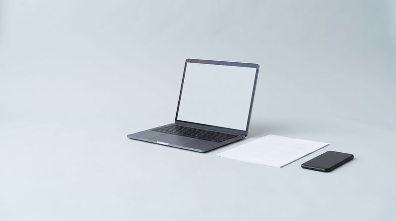 A dark laptop with blank white screen, a white notebook, and a black smartphone arranged on a light gray surface.